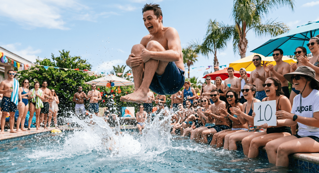 Man jumping into a pool during a lively pool party with friends and colorful umbrellas.