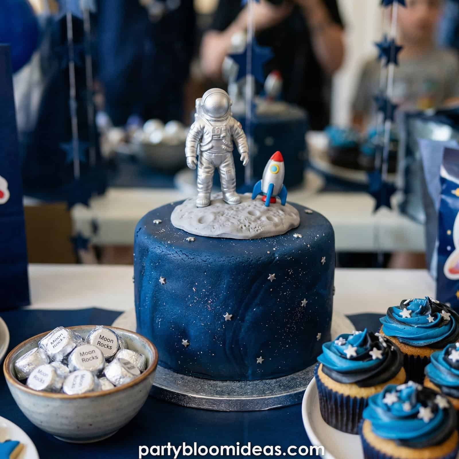 Space-themed birthday cake with astronaut and rocket toppers, surrounded by cupcakes with star decor.