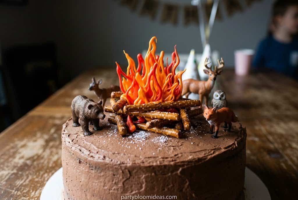 Camping-themed birthday cake decorated with pretzel logs, candy flames, and woodland animals