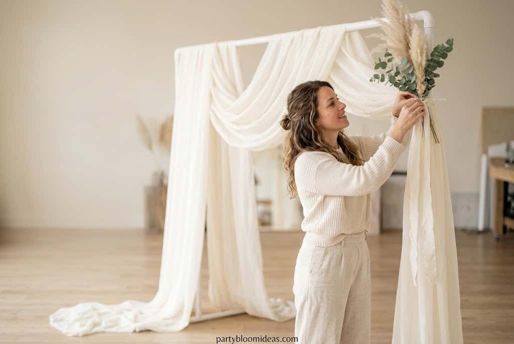 DIY wedding backdrop with flowing ivory fabric, pampas grass accents, eucalyptus sprigs, and elegant boho styling on a budget.